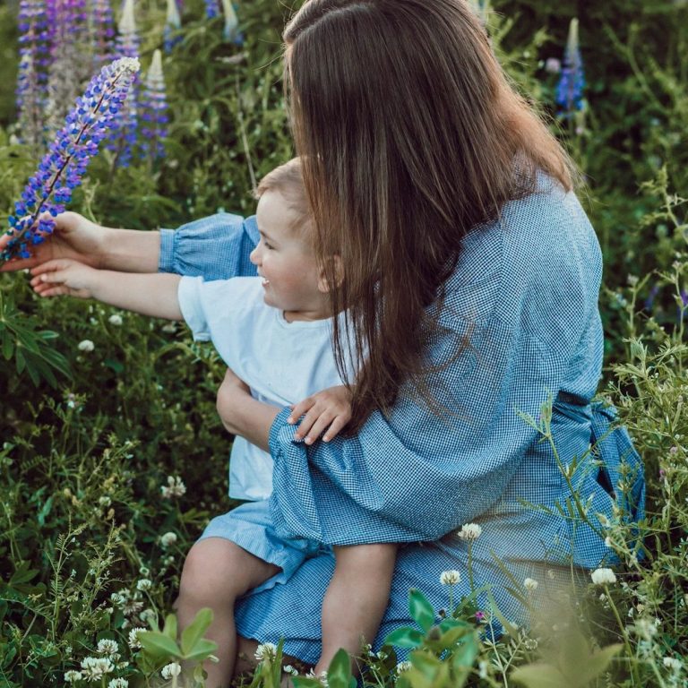 Eine Frau hält ein Kleinkind, das nach lila Blüten in einer Blumenwiese greift.
