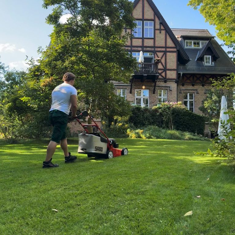 Mann mäht den Rasen vor einem großen, historischen Haus in einem grünen Garten.