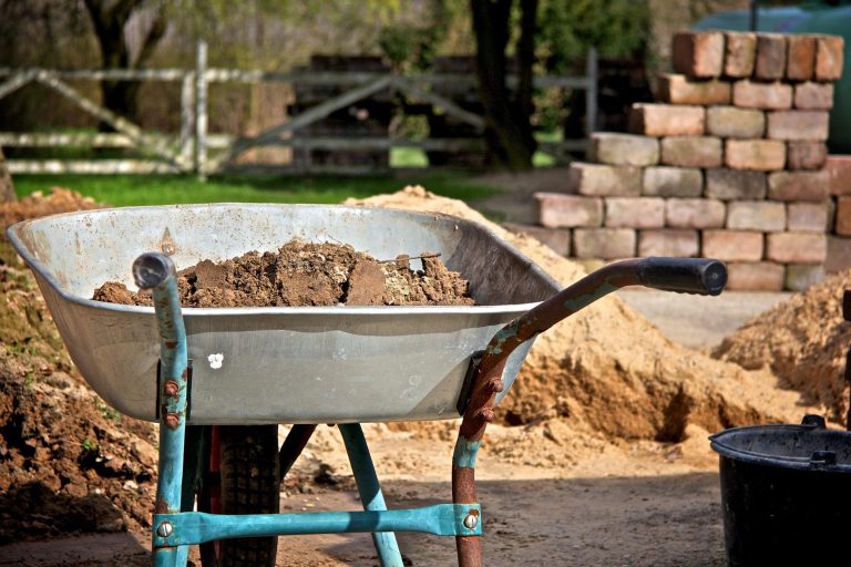 Schubkarre mit Erde, im Hintergrund ein Haufen Ziegelsteine und gelockerter Boden.
